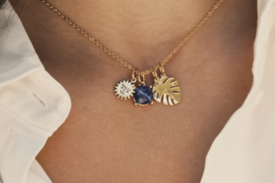 A person wearing an 18kt gold plated charm necklace with interchangeable charms, featuring a sun and a palm leaf design.